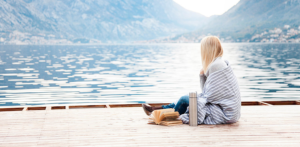 Junge Frau sitzt in ein Tuch gehuellt auf einem Bootssteeg am See